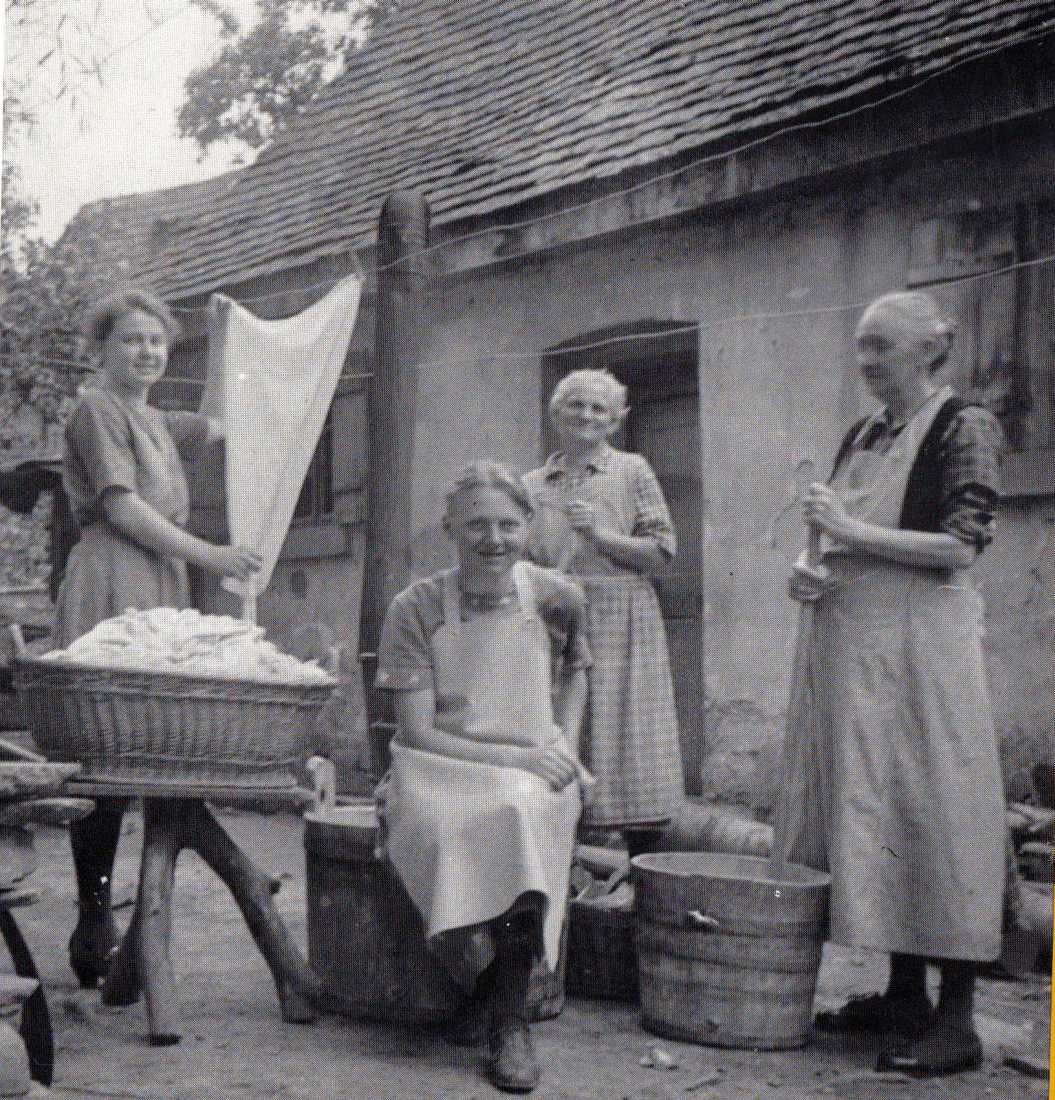  Vier Frauen vor dem jüdischen Badhaus. Eine Frau hängt die Wäsche auf. Zwei weitere Frauen stehen vor einem Waschbottich. Eine Frau sitzt auf dem Rand eines Wäschezubers. - Das Bild wird mit Klick vergrößert 