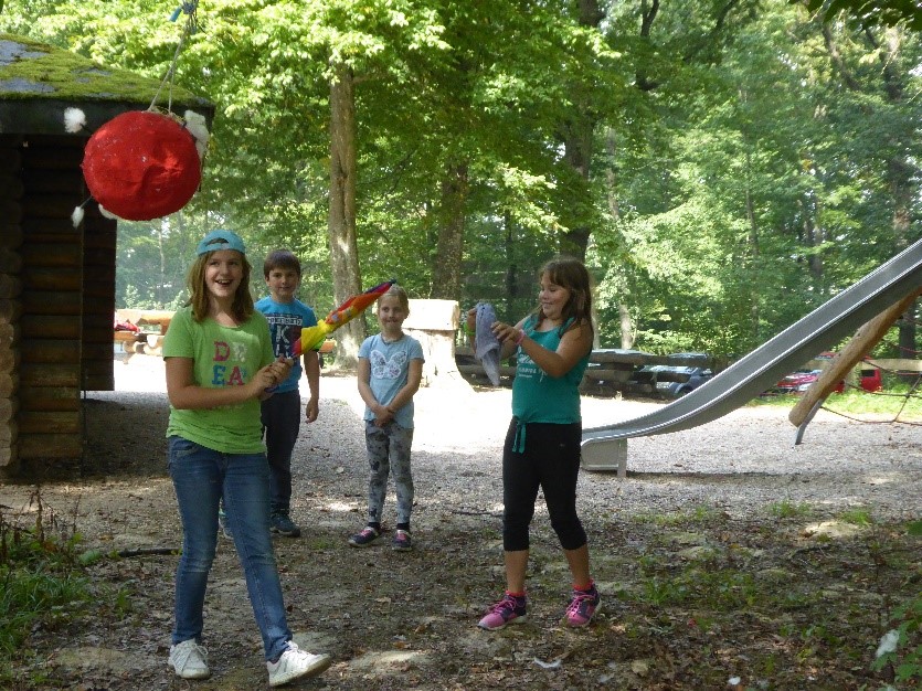  Im Wald: Ein Mädchen schlägt mit einem bunten REgenschirm auf die aufgehängte Corona-Pinata. Zwei weitere Mädchen und ein Junge stehen im Hintergrund und lachen. - Das Bild wird mit Klick vergrößert 