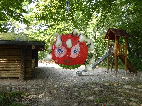  Im Wald: Die an einem Ast aufgehängte Corona-Pinata - sieht aus wie ein großer roter aus Pappmaché geformter Ballon. Verziert mit Gesicht und Wattebäuschen. - Das Bild wird mit Klick vergrößert.. 