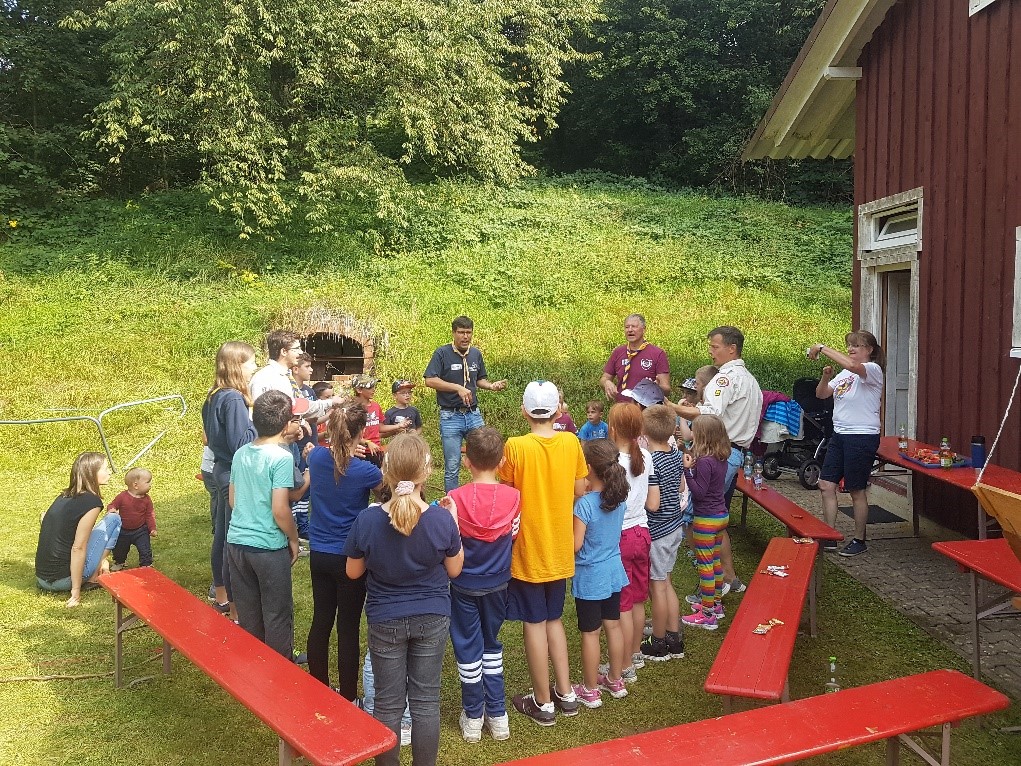  Eine große Gruppe Kinder im Alter von 7 bis 12 Jahren steht auf der Wiese neben der Pfadfinderhütte und hört zu, was die Betreuer sagen. - Das Bild wird mit Klick vergrößert 
